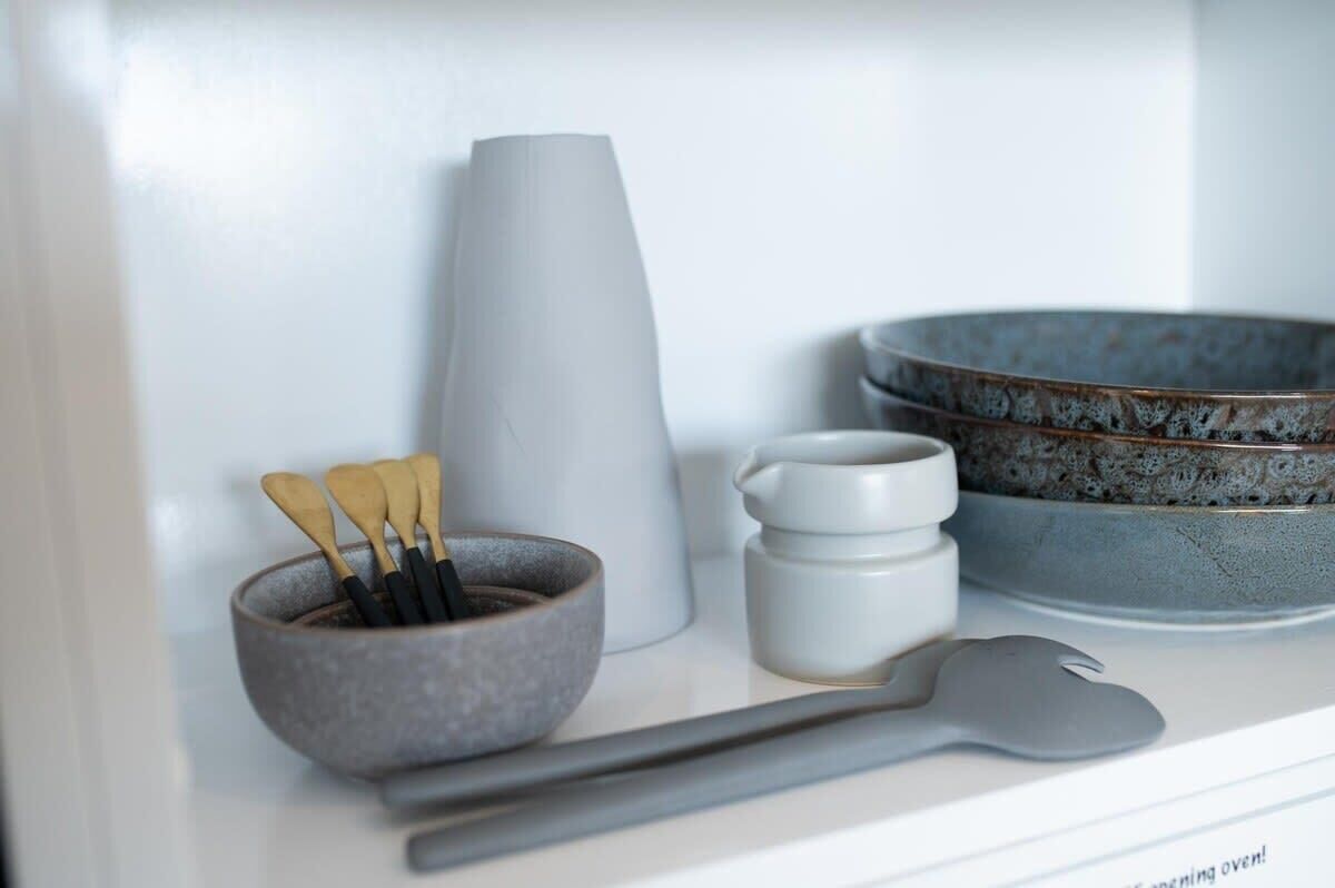 A kitchen counter with a bowl of utensils, a tall white container, a stack of plates, and a spoon.