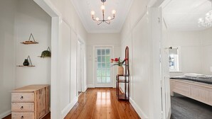 A hallway with wooden flooring, a white door, a bed with a grey bedspread, and a chandelier. - Bindi Cottage | Classic Charm, Short Stroll to CBD (Orange)