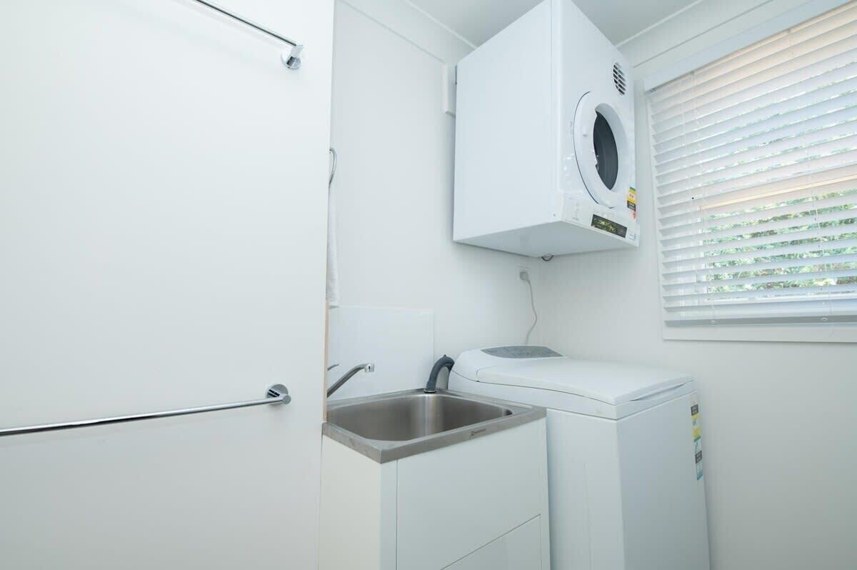 Une petite buanderie équipée d’un lave-linge, d’un évier et d’une fenêtre avec des stores.