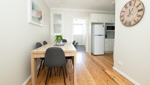 A dining area with a wooden table and chairs, a clock on the wall, and a kitchen in the background. - Sweet Caroline. Sunroom & Garden. Orange. (Orange)