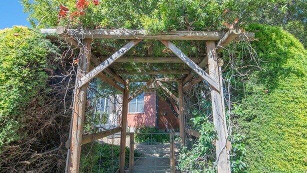 A wooden archway with a gate, surrounded by dense foliage and a building in the background. - Sweet Caroline. Sunroom & Garden. Orange. (Orange)