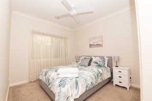 A bedroom with a bed, a nightstand, a ceiling fan, a window with sheer curtains, and a painting on the wall.