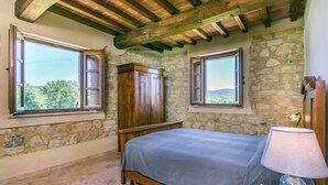 Quarto com paredes de pedra, vigas de madeira, cama, duas janelas e uma mesa de cabeceira com abajur.