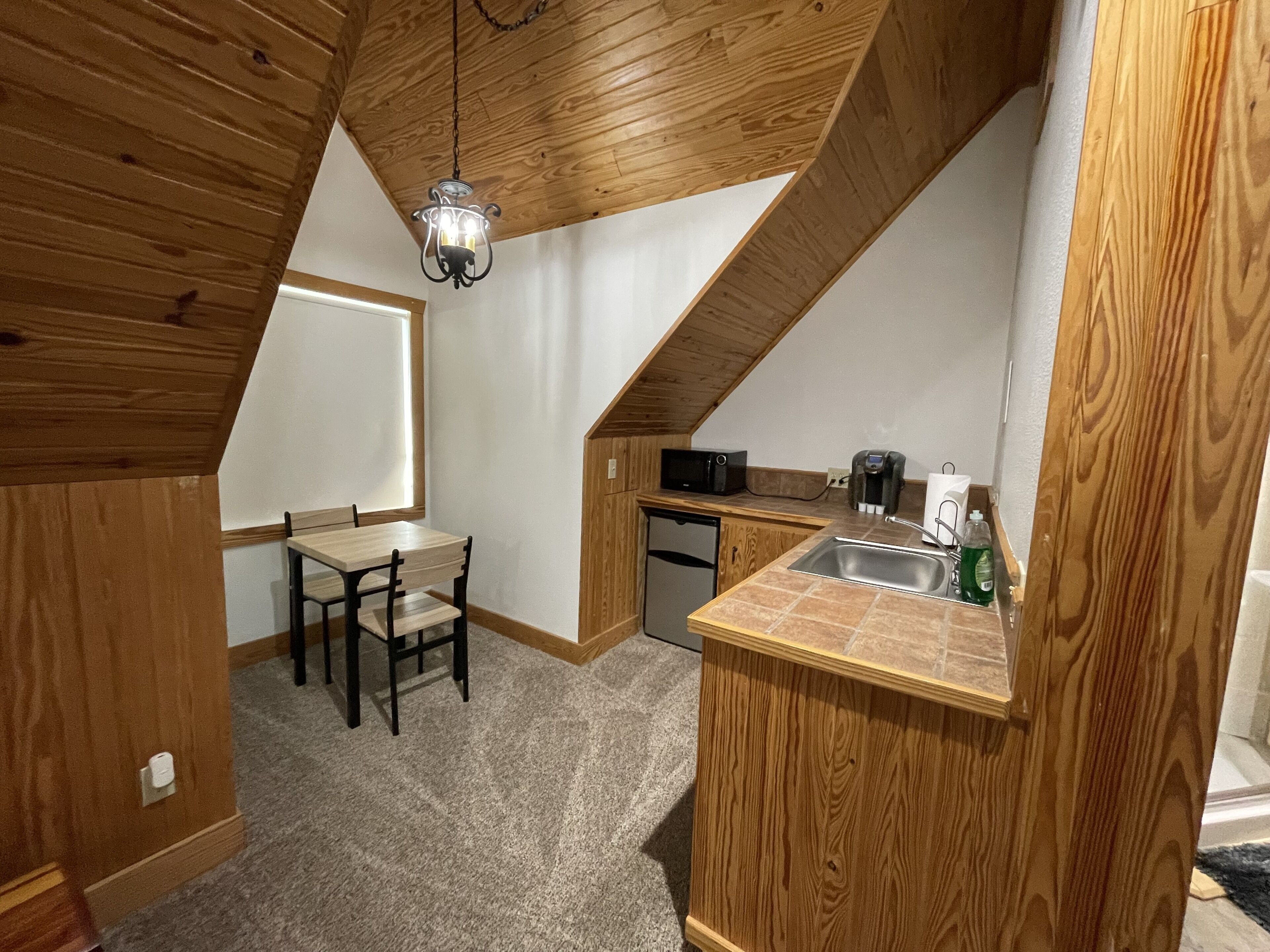 A compact kitchen with a wooden ceiling, a dining table, chairs, a microwave, a sink, and a small refrigerator.