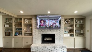 Een woonkamer met een witte bakstenen openhaard, een flatscreen televisie die erboven bevestigd is, en ingebouwde planken met diverse decoratieve objecten en boeken.