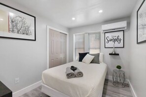 Un dormitorio con cama, una puerta, una ventana, aire acondicionado de pared y un cuadro enmarcado.