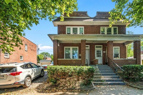 A two-story brick house with a front porch, a car parked in front, and a staircase leading to the entrance.