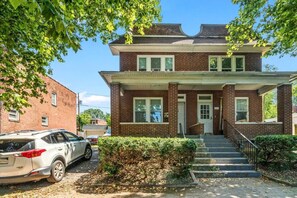 A two-story brick house with a front porch, a car parked in front, and a staircase leading to the entrance.