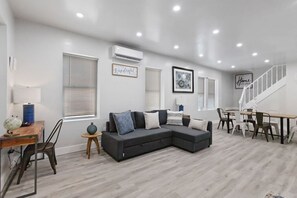 A modern living room with a sectional sofa, a dining table with chairs, and a staircase.