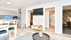 Una sala de estar moderna con comedor, una cocina de armarios blancos, un televisor de pantalla plana en la pared y una pequeña mesa con una planta.