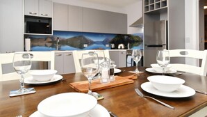 Un comedor con una mesa de madera dispuesta para cuatro, platos, tazones y copas de vino blancos. Hay saleros y pimenteros sobre la mesa. Al fondo, se ve una cocina con electrodomésticos modernos y un cuadro con un paisaje.