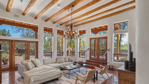 A spacious living room with a large sectional sofa, a coffee table, and two wicker chairs. The room features wooden beams on the ceiling, large windows, and a view of the outdoors. - Summer Pool, Hot Tub, Stunning Luxury (Santa Fe)