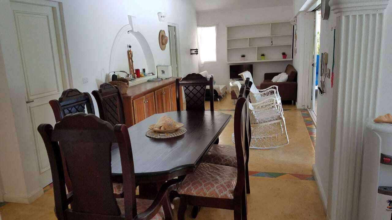 Un comedor con una mesa y sillas de madera, una cocina al fondo y un refrigerador blanco.