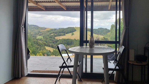 Quarto com uma grande janela com vista para uma paisagem montanhosa. Há uma mesa redonda com cadeiras e um vaso sobre ela.