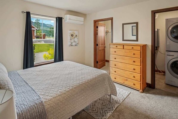 Un dormitorio con cama, cómoda de madera, lavadora y ventana con cortinas.