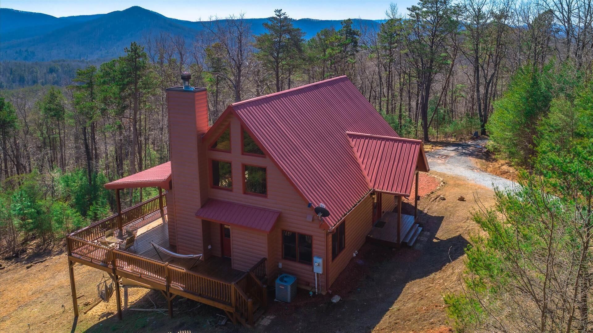 Una cabaña con techo rojo, una terraza y una chimenea, rodeada de bosque y montañas.