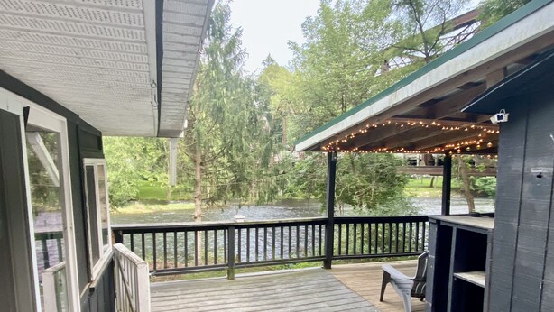 Une terrasse en bois avec une balustrade donnant sur une rivière, un espace salon couvert agrémenté de guirlandes lumineuses, et une vue sur les arbres et la verdure.