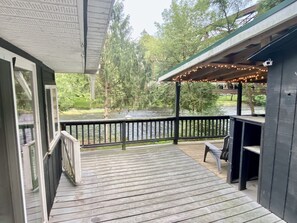 A wooden deck with a railing overlooking a river, a covered seating area with string lights, and a view of trees and greenery. - Waterfront Cabin Walkable to Everything! (Waterville)