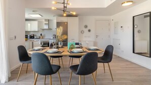 Un comedor moderno con una mesa redonda dispuesta para cuatro personas, rodeada de sillas oscuras con patas de madera. Un candelabro único con múltiples bombillas cuelga sobre la mesa. La habitación tiene un diseño minimalista con paredes de colores claros y un espejo grande en una pared.