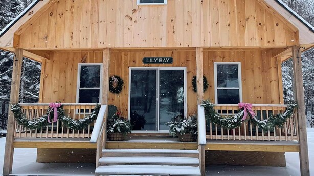 A wooden cabin with a porch, named "Lily Bay," featuring a front door, two windows, and a sign above the door. - Baker cabin|Downtown Greenville-Moosehead Lake|WIFI|Pets Ok|2 cabins available (Greenville)