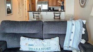 Un salon avec un canapé gris, un coussin décoratif et une vue sur la cuisine.