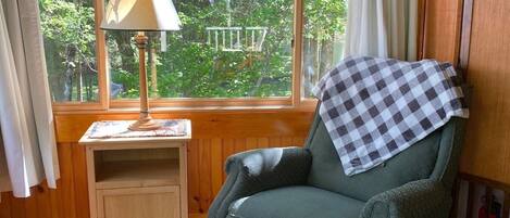 A cozy room with a green armchair, a small wooden nightstand, a lamp, and a view of trees through a large window.