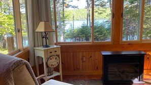 Una habitación acogedora con cama, una mesita de noche con lámpara, chimenea y ventana con vistas a árboles y agua.