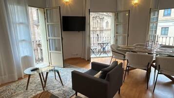 Una sala de estar moderna con un televisor de pantalla plana, un comedor con una mesa de vidrio y vistas a un patio a través de puertas abiertas.