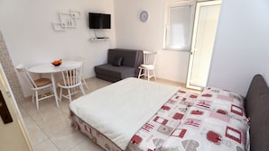 Ein Schlafzimmer mit einem Bett, einem kleinen Tisch mit Stühlen, einem Sofa, einem Fernseher und einem Fenster mit Jalousien.