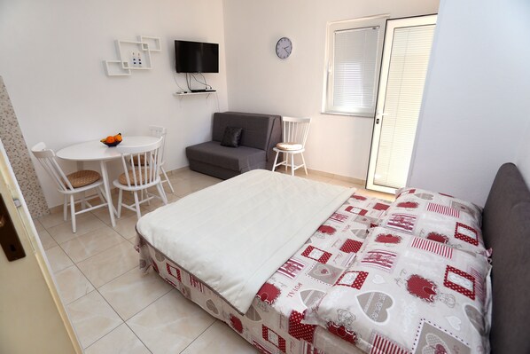 A bedroom with a bed, a small table with chairs, a sofa, a TV, and a window with blinds.