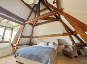 A bedroom with a sloped ceiling, exposed wooden beams, a bed with a patterned bedspread, a bedside chair, and a window with a view of the outside.