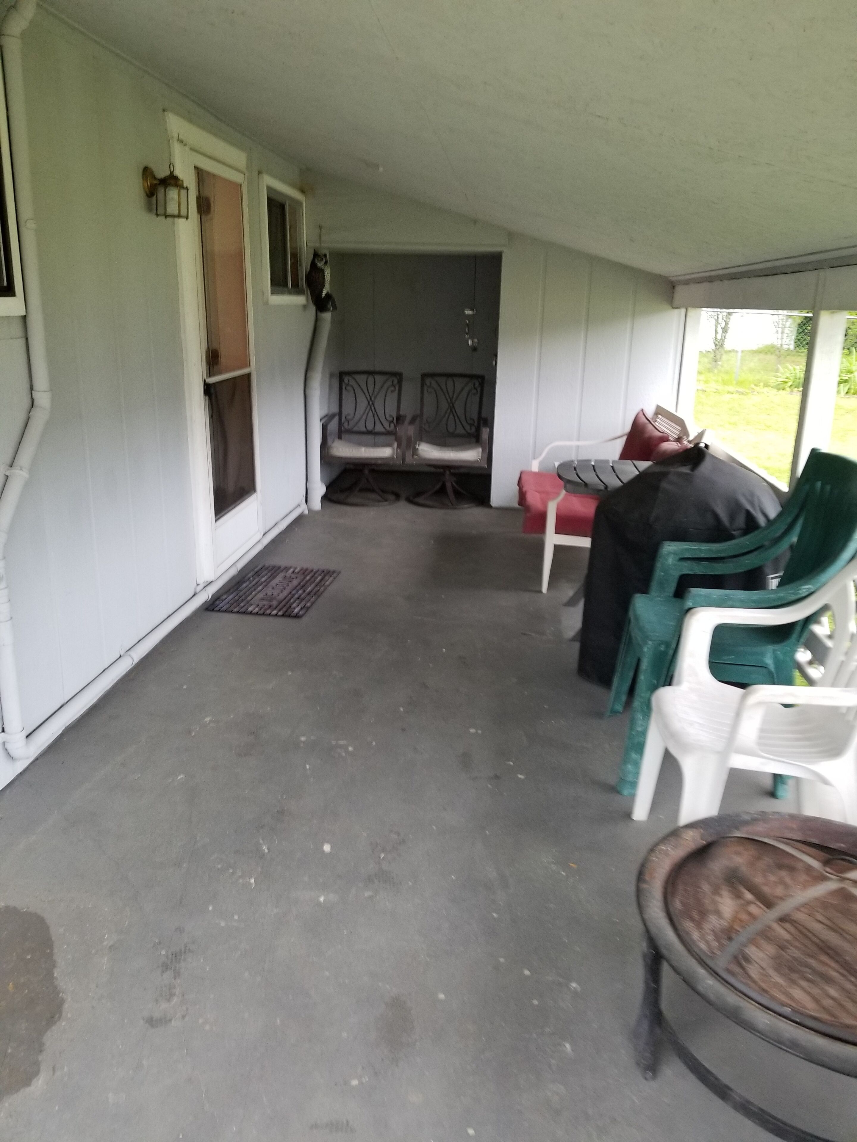 Eine überdachte Veranda mit zwei Metallstühlen, einem Tisch mit rotem Kissen und einem grünen Plastikstuhl.