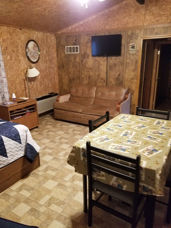 A cozy room with a brown leather sofa, a wooden table with a patterned tablecloth, a small wooden cabinet, and a television mounted on the wall.