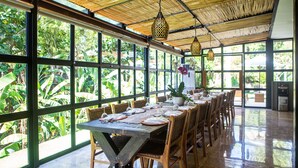 A dining area with a long table set for multiple people, surrounded by large windows offering a view of lush greenery. - Sunset Paradise Nusa Dua (Nusa Dua)