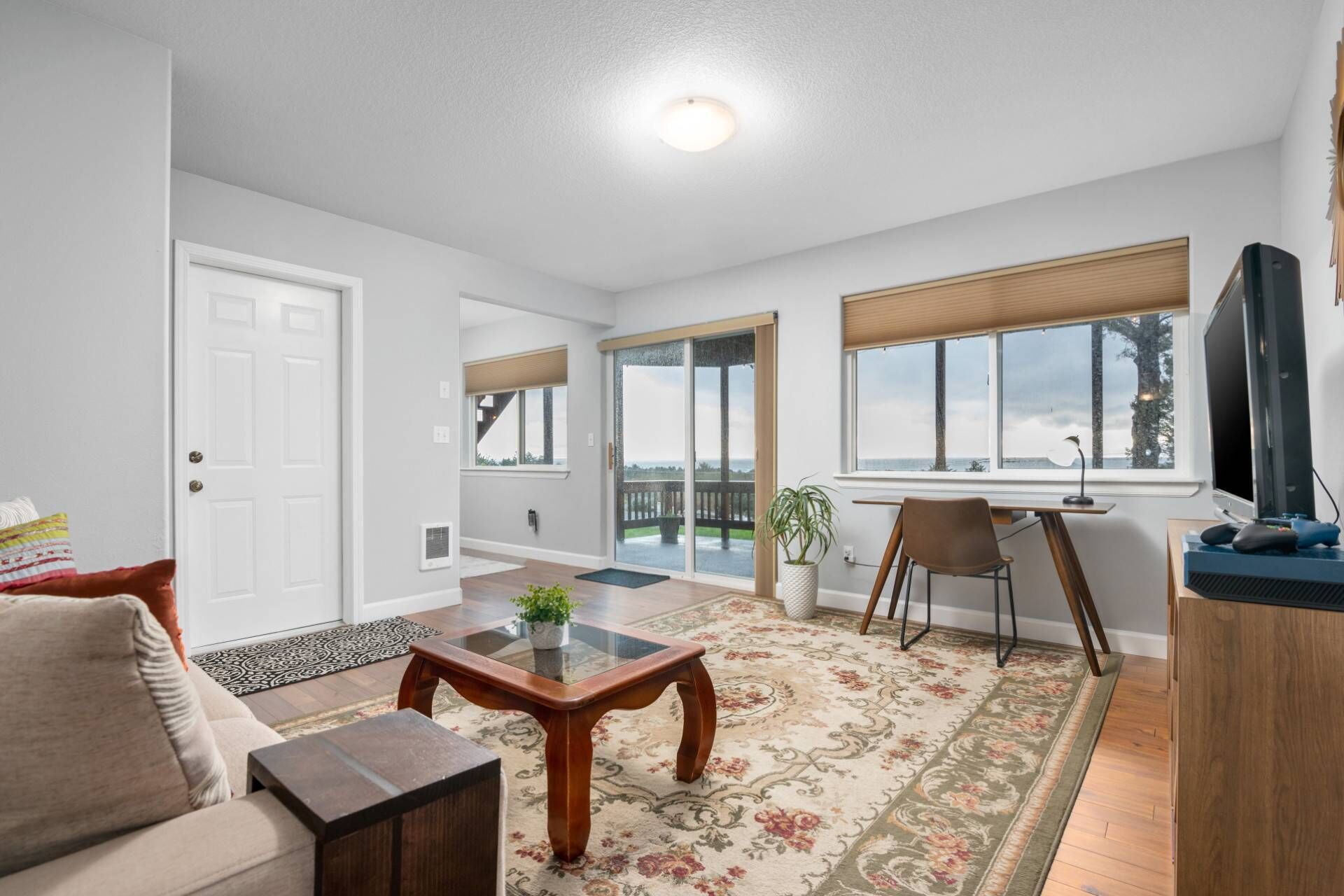 A spacious living room with a sofa, coffee table, and a TV on a wooden stand. There is a large window with a view of the outdoors.