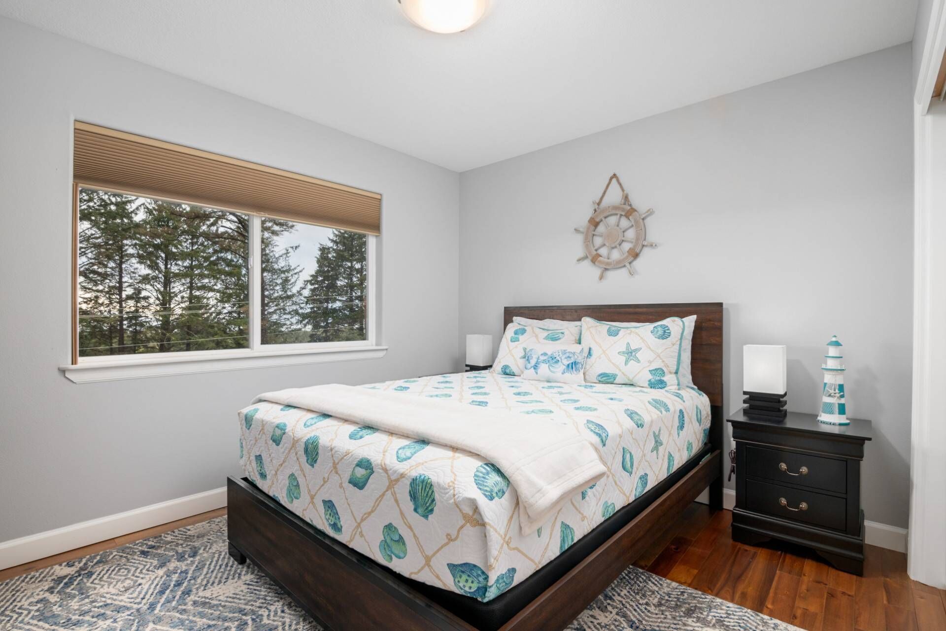 A bedroom with a bed, a nightstand, a wall-mounted ship's wheel, and a window with a view of trees.