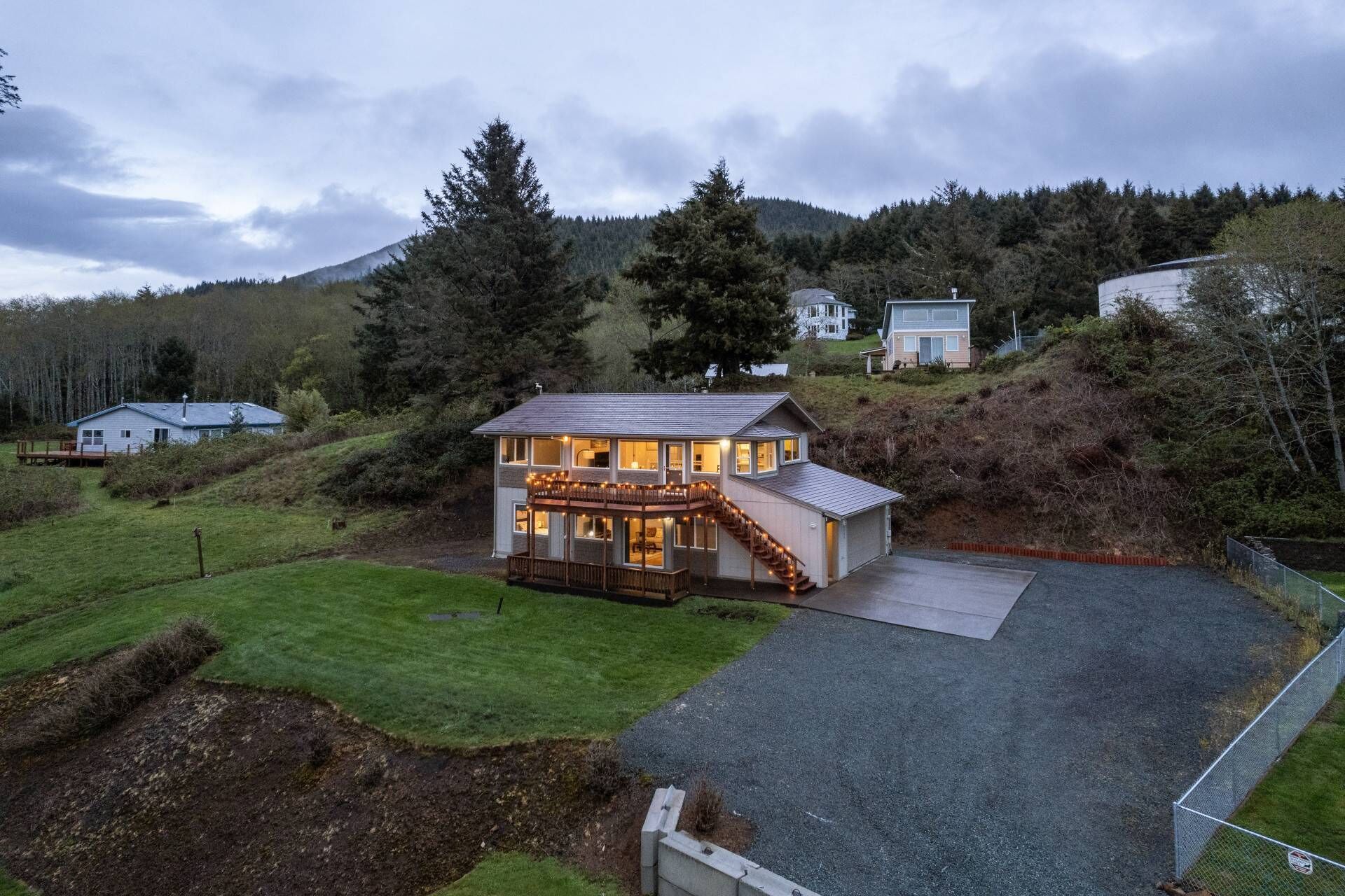 A two-story house with a deck and a covered driveway, surrounded by a forested hillside.