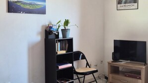 A room with a bookshelf, a chair, a television on a wooden stand, and two framed pictures on the wall. - T2 Escale sudiste, south of Reunion Island (Saint-Louis)