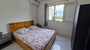 A bedroom with a bed, a ceiling fan, an air conditioning unit, a window with curtains, and a refrigerator. - T2 Escale sudiste, south of Reunion Island (Saint-Louis)