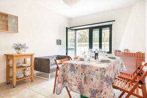 A dining area with a table set for four, a wooden side table with dishes, a sofa, and a view through a large window.