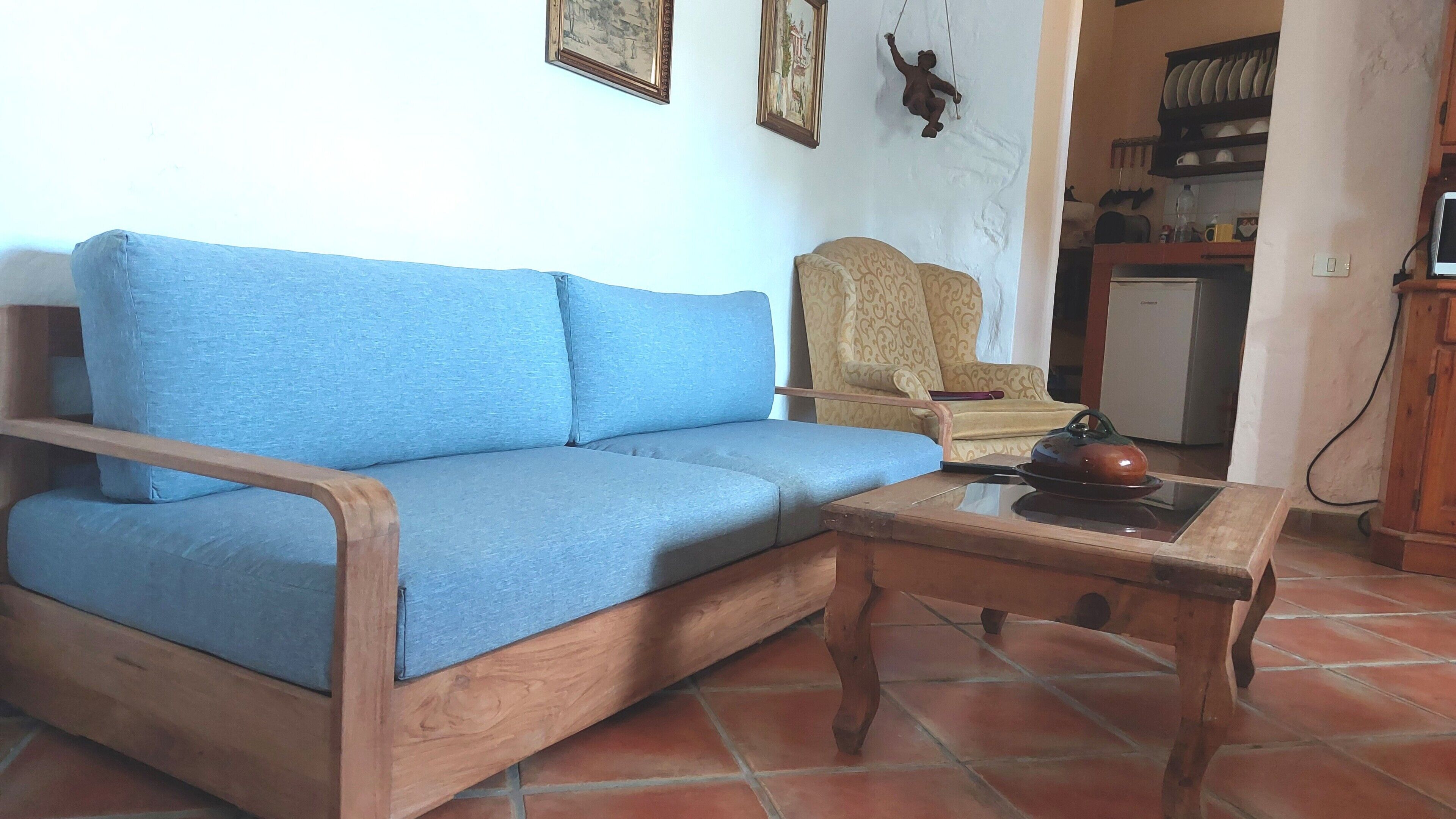 Um sofá azul, uma mesa de centro de madeira, uma poltrona estampada e uma área de cozinha com geladeira e louças.