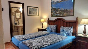 A bedroom with two beds, a nightstand, a lamp, a painting, and a bathroom visible through an open door.