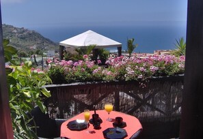 Ein Tisch mit rotem Tischtuch, ein Glas Orangensaft und eine schwarze Tasse, mit Blick auf eine Küstenlandschaft mit rosa Blumen und einem weißen Sonnenschirm.