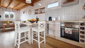 Une cuisine avec des poutres en bois, des meubles blancs, un poêle et un coin repas avec des chaises blanches.