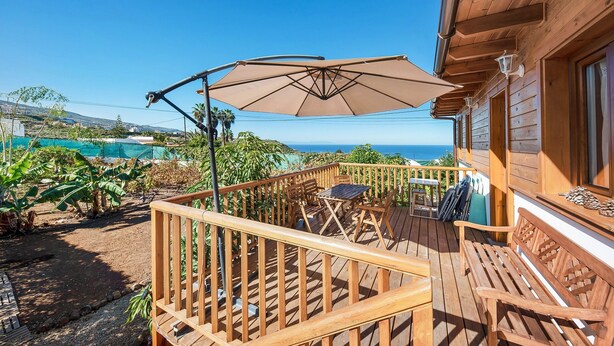 Une terrasse en bois avec une table et des chaises, un grand parasol et une vue sur l’océan.