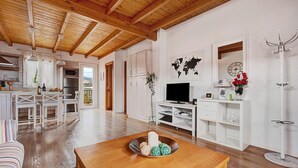 Un vaste espace de vie doté d’un plafond en bois, un coin repas avec des meubles blancs, une cuisine avec des placards blancs et un meuble de rangement blanc intégrant un téléviseur à écran plat.