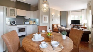 Een moderne keuken met witte kasten, een eettafel gedekt met witte servies en een zithoek met een bruine bank en een televisie.