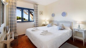 Un dormitorio con una cama, dos mesitas de noche, un ventanal con cortinas, vista a árboles y un pequeño cuadro en la pared.
