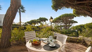 A round table with chairs, a basket of fruit, a teapot, and cups on a patio with trees and a lamp in the background.