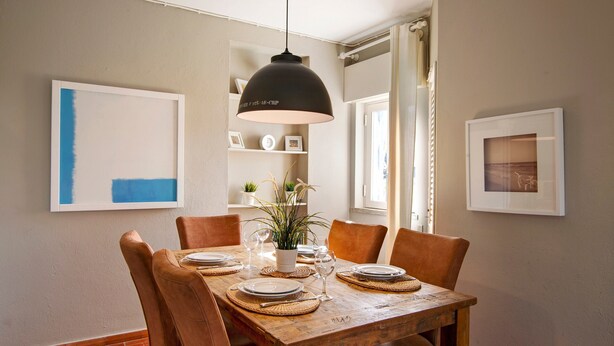 A dining area with a wooden table set for four, surrounded by chairs, with a pendant light above.